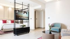 Modern hotel suite featuring a cosy seating area with a blue armchair, glass-top coffee table with fresh flowers, and a plush bed with red accent pillows—divided by a sleek black TV stand for a stylish open-plan layout - Golden Tulip, Lucknow