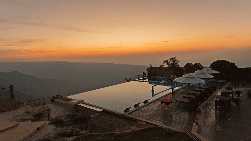Cliffside pool deck at Polo Resort Cherrapunji at twilight with infinity pool, white parasols, lounge seating and silhouette of valleys under orange sky.