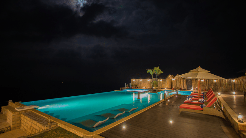 Illuminated infinity pool at night at Polo Resort Cherrapunji with red loungers, bamboo screens, wooden deck and a cloudy moonlit sky overhead.