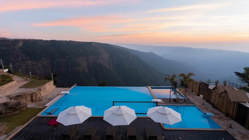 Polo Resort Cherrapunji infinity pool at sunset with three white parasols, sun loungers, wood decking and dramatic canyon and mountain views beyond.
