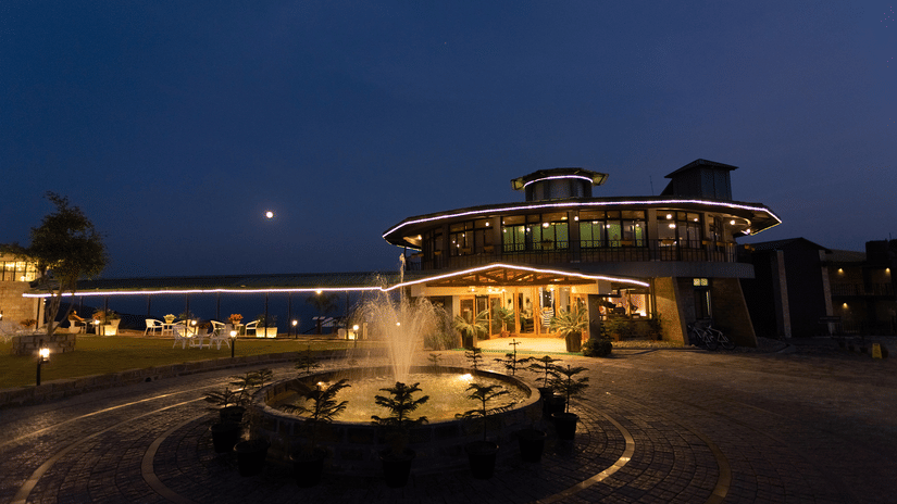 Night facade of Polo Resort Cherrapunji main building outlined in LED lights, with a circular driveway, illuminated fountain and potted conifers in front.
