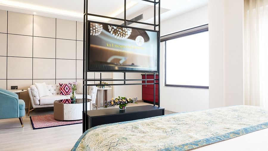 Spacious hotel suite with a bed in the foreground, large TV mounted on a modern frame, and a stylish sitting area with plush chairs, a rug, and vibrant cushions - Golden Tulip, Lucknow