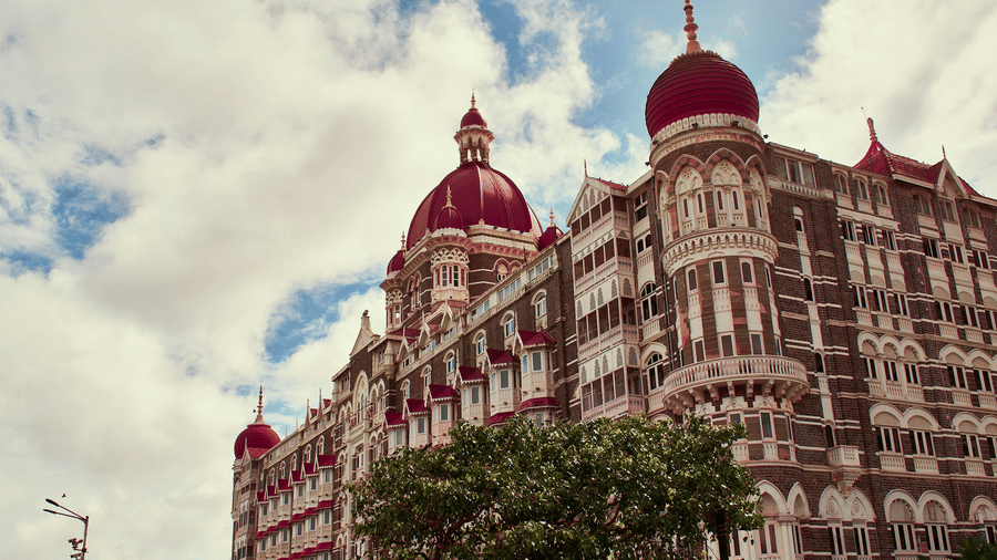 The large Taj Hotel building with domes and arched windows, with trees in front and clouds in the background.
