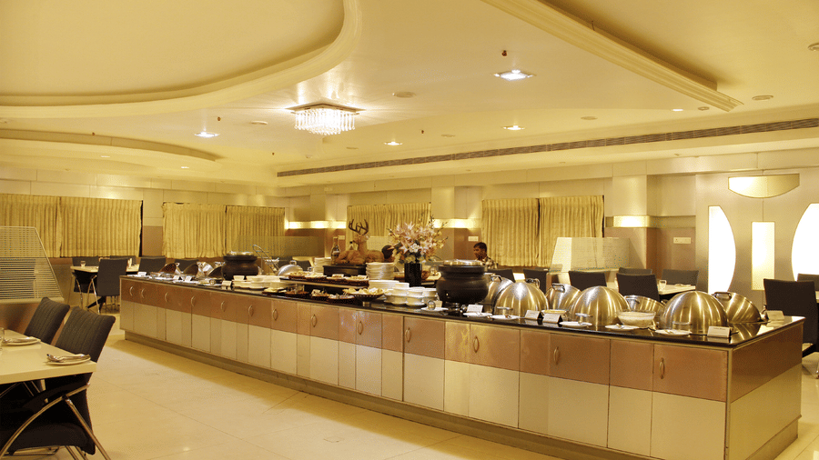 A buffet counter at DSF Grand Plaza, Tuticorin with silver chafing dishes, a floral arrangement, and a deer figurine, set against curtained windows and dining tables.
