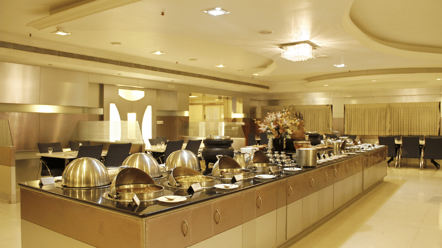 A buffet counter at DSF Grand Plaza, Tuticorin with open chafing dishes revealing food, place cards, a floral arrangement, and dining tables in the background.