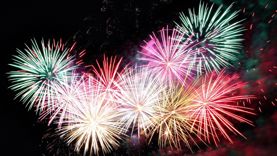 Multi-coloured fireworks exploding brightly over a dark pier structure and water at night.
