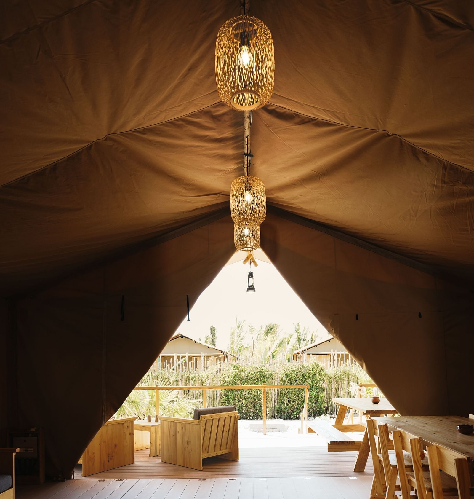 Sneak peak of the area outside the tent featuring chairs in the balcony and cottages at short distance. Inside the text of WoCAL there are table and chairs and a lamp hanging from the ceiling. 