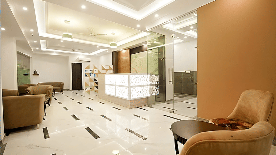 A waiting area at Hotel Apple Park Delhi located near a glass lift. The space includes a tan sofa, a wall-mounted television, and a small coffee table.