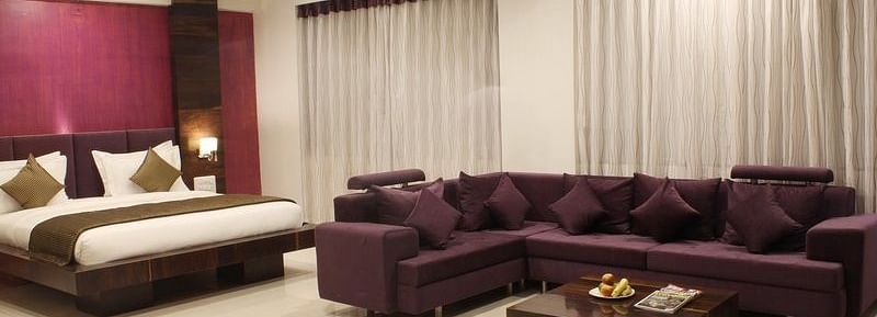 A spacious hotel suite features a king size bed, a comfortable brown sofa set around a coffee table, and large windows with sheer curtains.