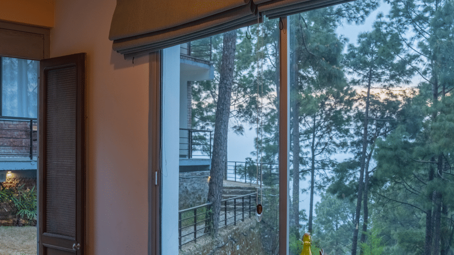 A dining table with chairs is positioned next to a large window at BluSalzz Escapade Resort, Lansdowne. Trees are visible outside the window.