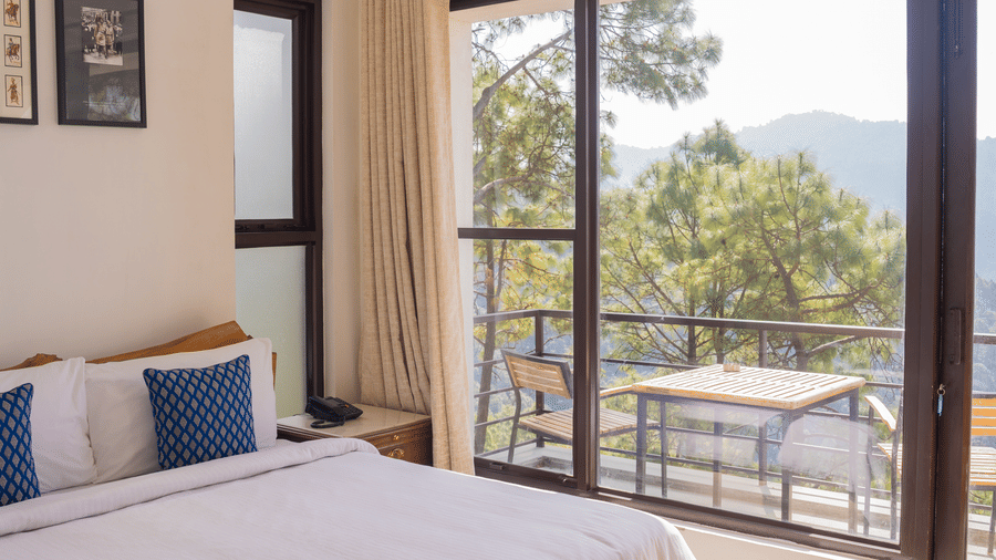 Sunlight streams into the Sun Terrace Room at BluSalzz Escapade Resort, Lansdowne, illuminating a bed and a bench. Large glass windows offer a view of the outdoors.