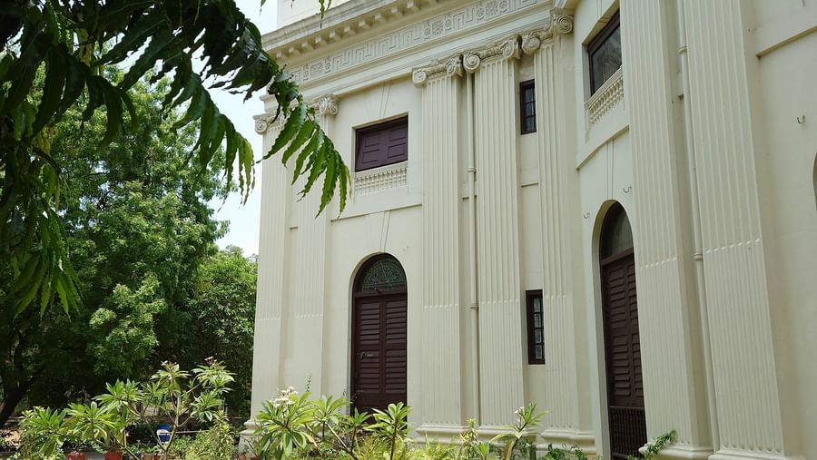 A white heritage-style building with columns and arched windows, surrounded by plants and trees.