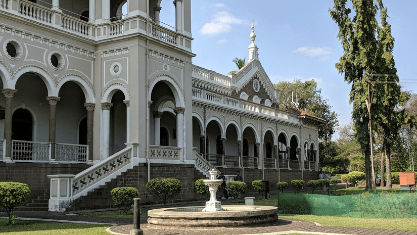 A facade view of Aga Khan Palace showcases arched corridors, upper balconies and an open lawn with a sculpted fountain.