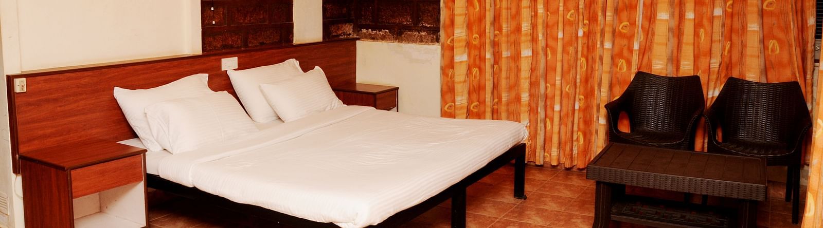 A spacious room at Wind Chalet Resort with a large bed and pillows against a wooden headboard, a bedside table, and a table with a pair of chairs at the foot of the bed, with curtains covering the windows.
