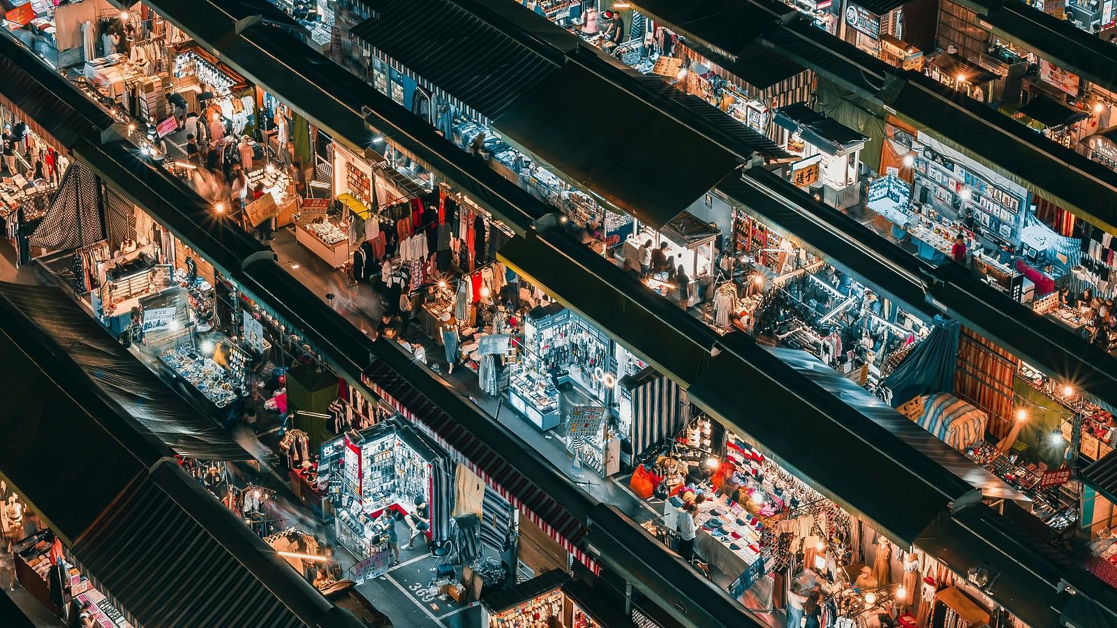 An aerial view of a bustling night market, rows of stalls illuminated with warm light, creating a textured, vibrant pattern.