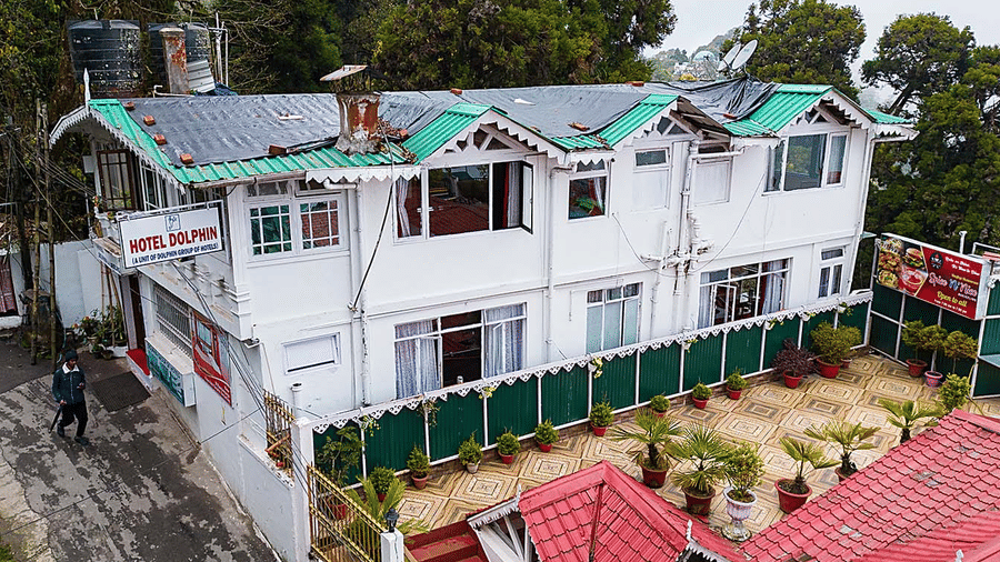 An aerial view of a multi-level white hotel building with green and red roofs nestled among trees on a hillside at Hotel Dolphin Darjeeling.
