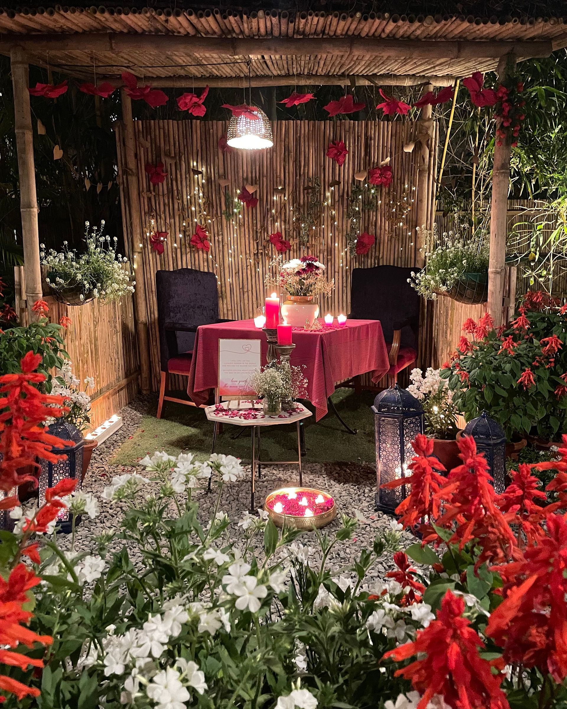 Alt-text: Romantic outdoor dining setup with a red-themed table for two, surrounded by flowers, candles, and warm lighting under a wooden canopy - Karma Chalets