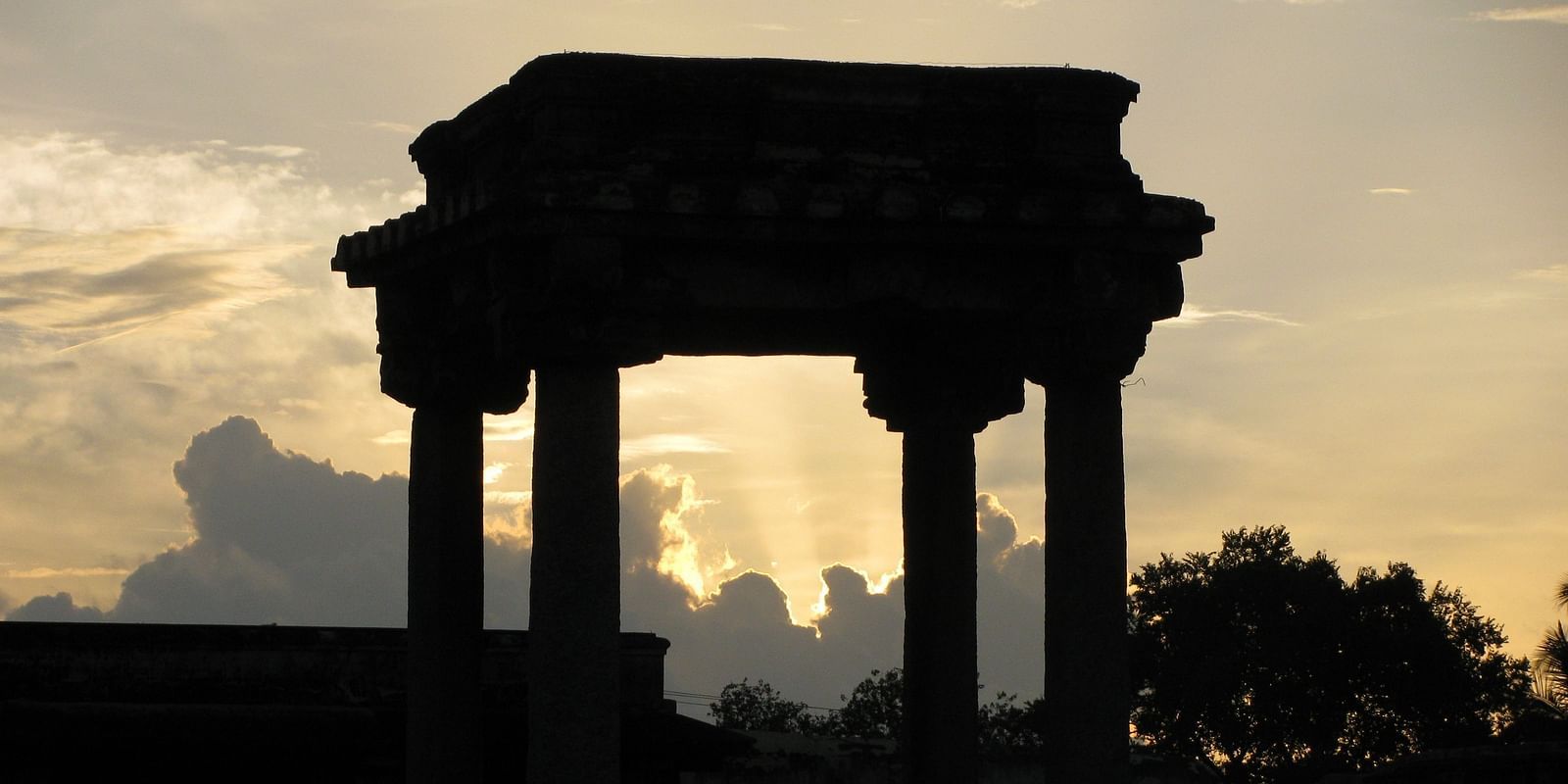 An overview of ancient columns in Belur with the sun's rays behind the clouds in the background.