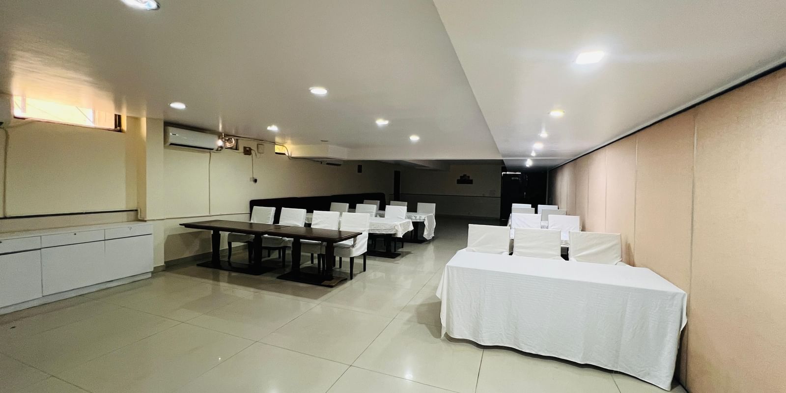  A hotel conference room with long tables and white chairs arranged for an event, featuring modern lighting and light-colored walls. | Hotel Aadhar