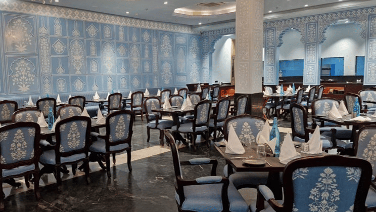 An elegant dining room at Beelwa Palace, Jaipur displays classic blue upholstered chairs and sparkling chandeliers.