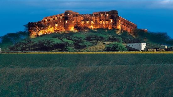 Hill Fort-Kesroli featuring a structure at the top of a hill illuminated with lights. The fort is surrounded by trees, plants, and field.