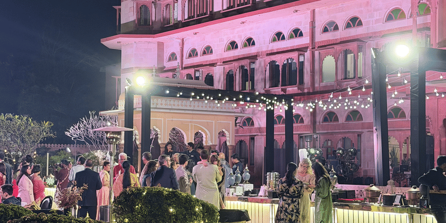 A night view of a wedding event on the lawn with the grand heritage facade of Umaid Palace brightly lit in pink and white light.