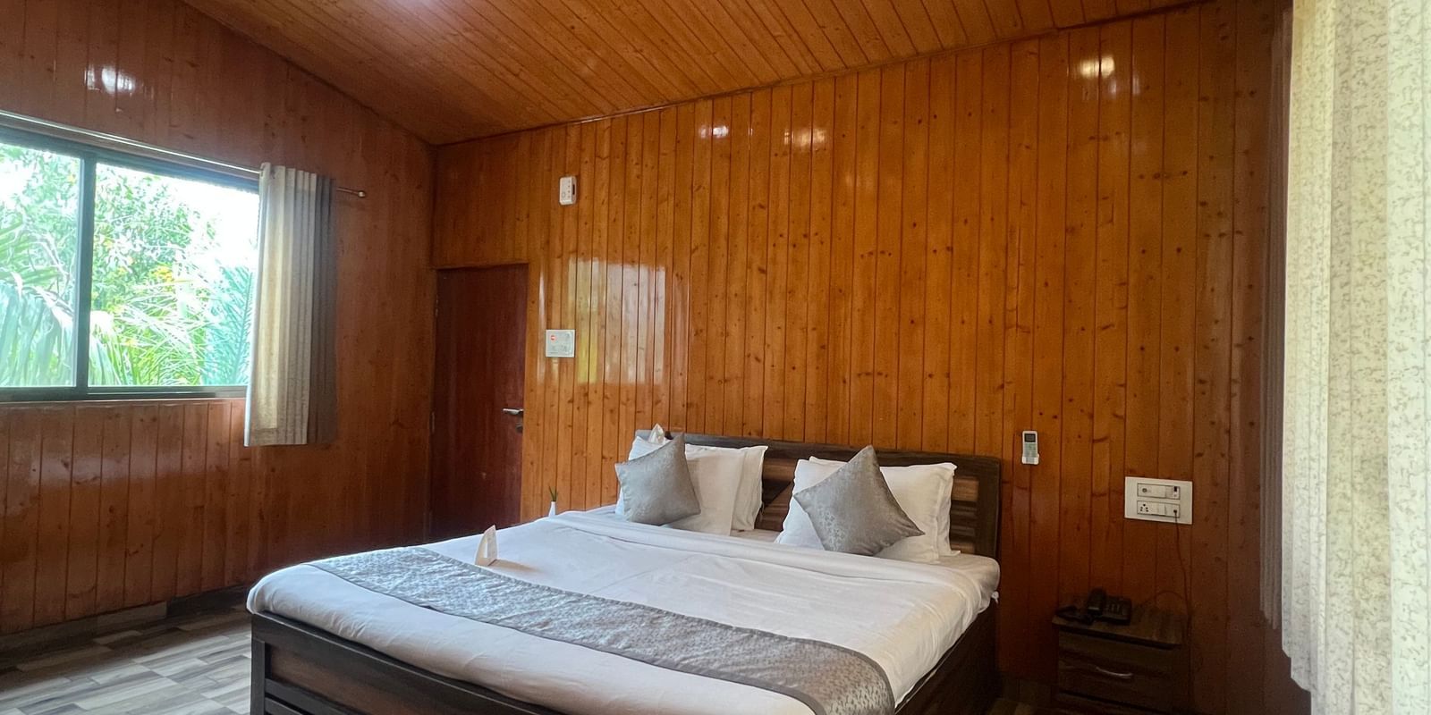 Wide view of the Daksh Elite Cottage, highlighting its wooden interiors, a window with curtains opened, and its centrally placed double bed, at Daksh The Valley Resort, Saputara.