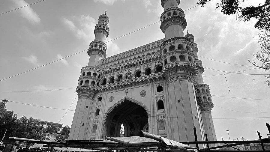 Black and white, low-angle view of Charminar, a grand archway with four minarets, towering over a bustling market area.