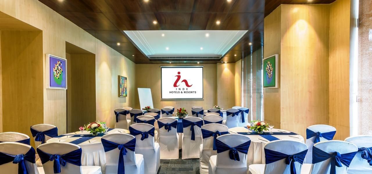 Meeting room at Inde Reva Resorts Rishhikesh, with white chairs tied with navy blue sashes, round tables decorated with flower arrangements, and a projector screen at the front.