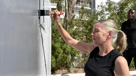 A woman in a black sleeveless top signs or writes on a large gray board outdoors with buildings and greenery in the background, while another person watches nearby.