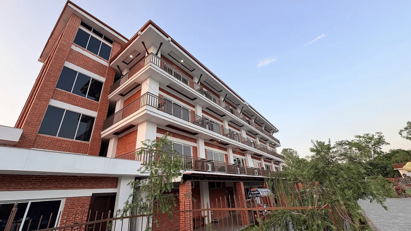 Red-brick facade with white columns, railed balconies and a landscaped forecourt with young trees and paved paths at Ila Comfort, Harkapur.