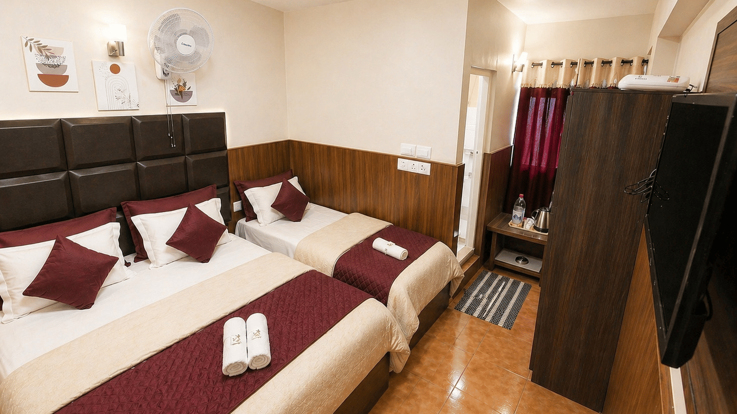 A double bed next to a single bed, with a wall-mounted TV and a wardrobe in the Deluxe Triple Room at BN Tourist Home, Ooty.