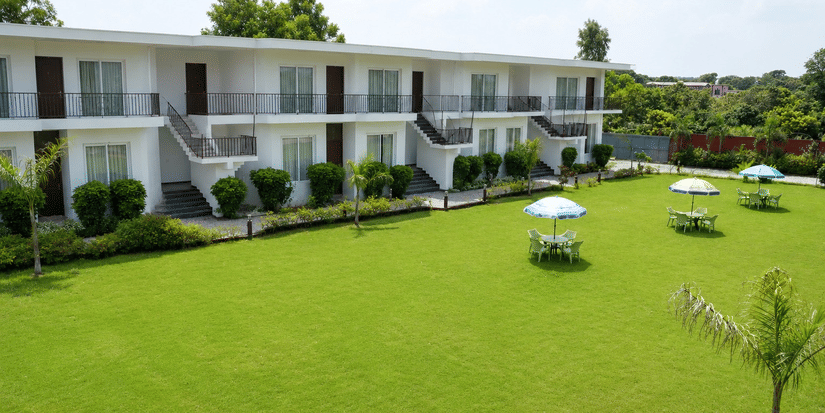 Exterior of a white 2-storey resort building with balcony railings and rooms facing a large manicured green lawn with shaded outdoor seating tables - Five Elements Hotels, Corbett Crown Resort, Jim Corbett