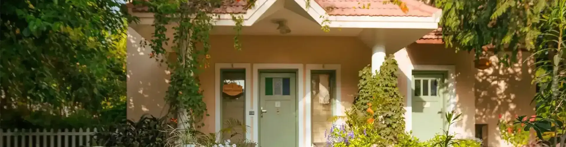 A small house with a white door, porch, and red roof, surrounded by lush greenery and a white picket fence.