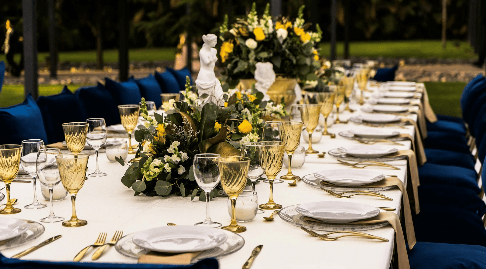 A rectangular table in an outdoor setting with cutleries arranged on it.