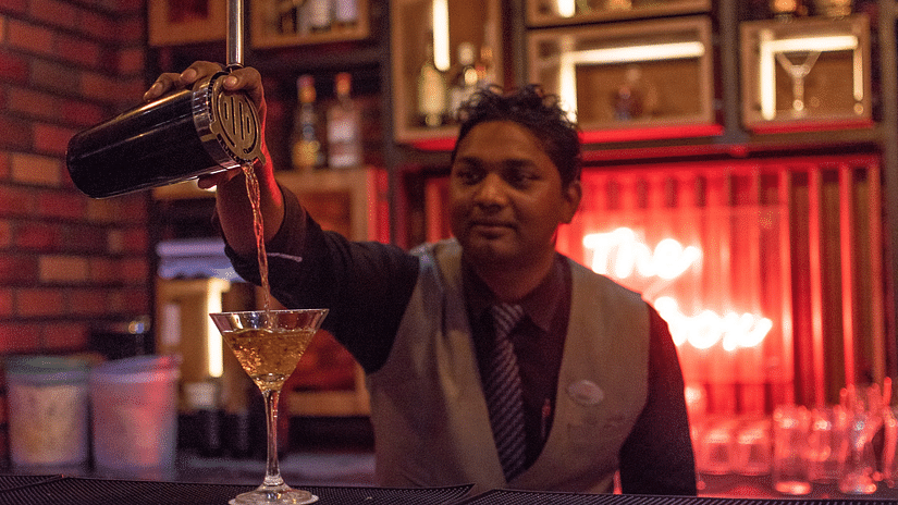 A man tilts a bottle, pouring a drink into a glass from a height - The Open Box - Wedlock Greens Hotel Resorts Dhanbad