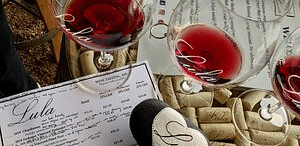Wine glasses filled with red wine rest on a table with menus, corks, and a heart-shaped object