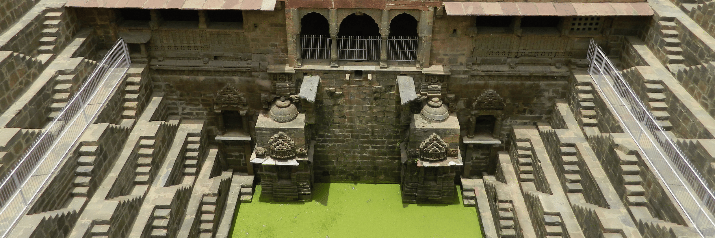 The vast, symmetrical stone steps of Chand Baori stepwell descend towards a pool of bright green water.