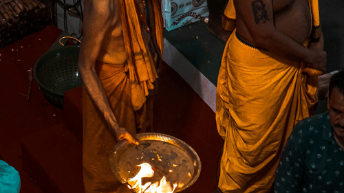 A person in traditional attire is performing a ritual with a lit oil lamp in a dimly lit indoor setting.