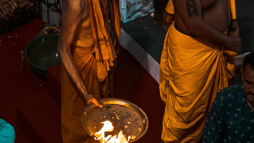 A person in traditional attire is performing a ritual with a lit oil lamp in a dimly lit indoor setting.