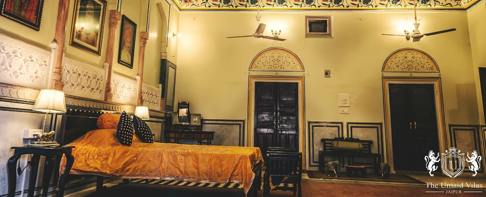 A spacious room at Umaid Vilas Jaipur with a patterned ceiling, multiple fans, and a bed with an orange throw.