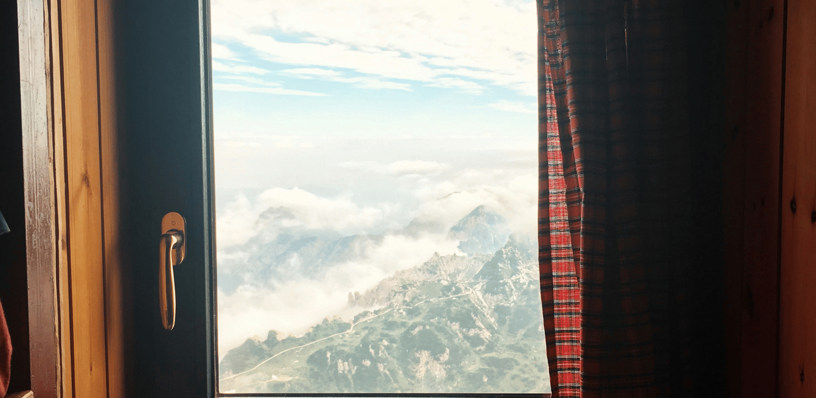 A window framed by dark wood, looking out onto a stunning vista of rolling, misty mountains under a bright, cloudy sky.