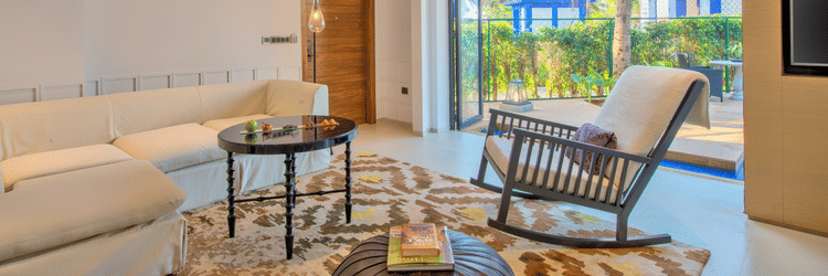 A living area of the room at Azaya Beach Resort Goa with a rocking chair, pouffe, table and couch facing the large door opening to the pool deck