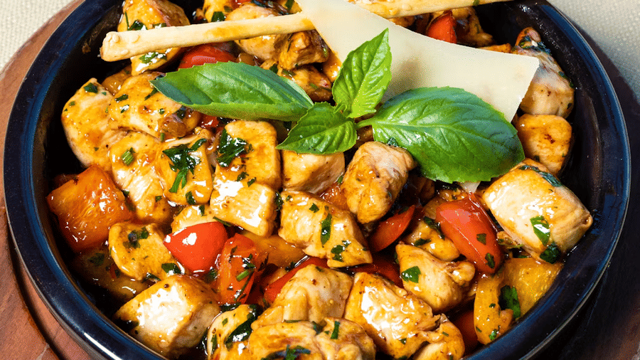 Bowl of glazed grilled chicken cubes with red peppers, chopped herbs, a basil sprig, a cheese shaving, and 2 sesame breadsticks on a wooden board.