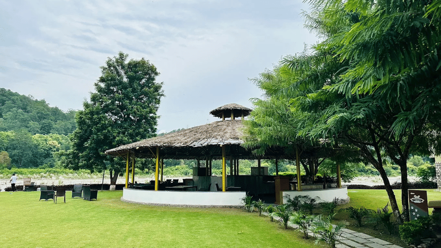 An open-air, thatched-roof dining pavilion sits on a grassy lawn with trees, overlooking a deck near a riverbank under a cloudy sky.