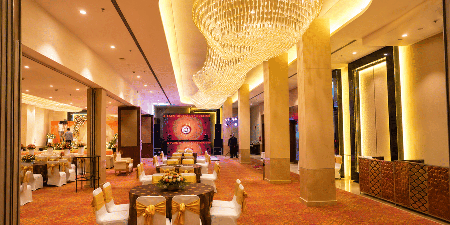 Spacious banquet hall with a chandelier ceiling, dining tables, and a stage area at Ambrosia Sarovar Portico, Haridwar.