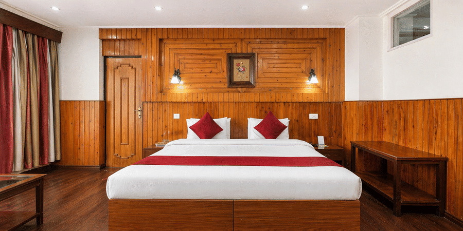 Front view of a room at Central Hotels featuring a king bed with red accent bedding wooden wall panels dark hardwood flooring warm wall lamps and red curtains on the window.