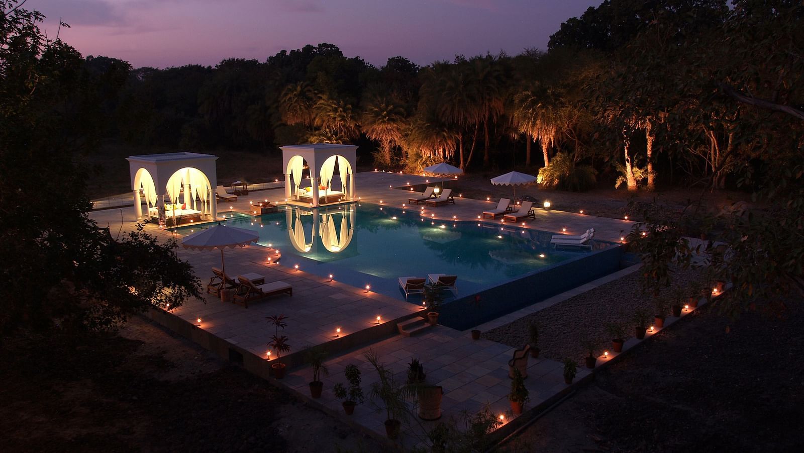 An illuminated swimming pool at night, surrounded by trees and structures with arched openings, creating a serene and inviting atmosphere- Shahpura Bagh.