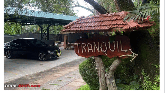 A Team-BHP travel article featuring a black sedan parked next to a rustic wooden welcome sign at the entrance of Tranquil Resort, Wayanad.