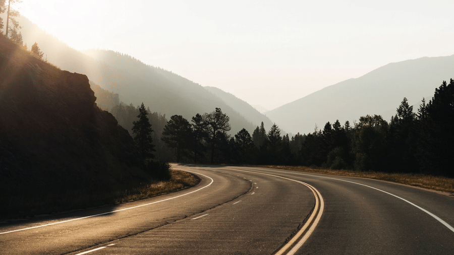 Empty curved mountain highway with double yellow lines winding through pine-forested hills with a glowing sunburst peeking over the ridge.